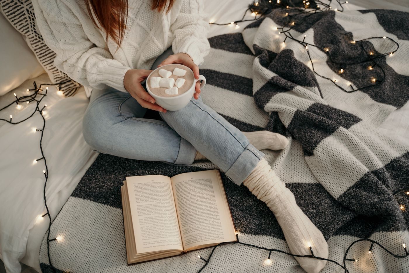 Person in wool socks with a mug of hot chocolate and a book on a blanket with fairy lights